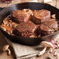 steaks and mushrooms cooking in a skillet on a wooden table next to sliced mushrooms