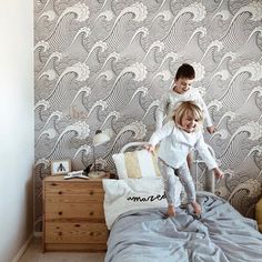 two children jumping on a bed in front of a wallpapered room with waves