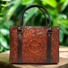 a brown tooled leather bag sitting on top of a wooden table next to green leaves