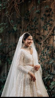 a woman wearing a wedding gown and veil standing in front of a wooden fence with greenery