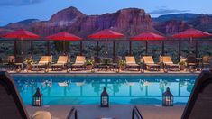 an outdoor pool with chairs, umbrellas and mountains in the background at night time