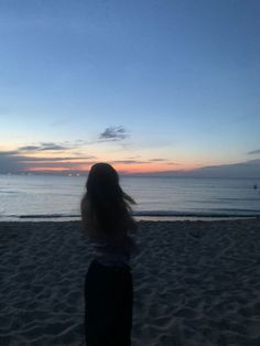 a woman standing on top of a sandy beach next to the ocean at sunset with her hair blowing in the wind