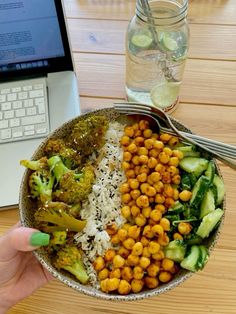 a person holding a bowl filled with vegetables and chickpeas next to a laptop