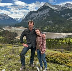 a man and woman standing on top of a grass covered hillside next to mountains with snow capped peaks in the background