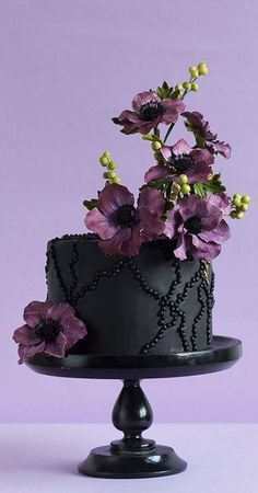 a black cake decorated with purple flowers on a stand in front of a purple background