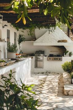 an outdoor kitchen with white walls and stone flooring is seen through the leaves of a tree