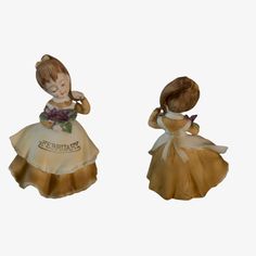 two figurines of girls dressed in yellow and brown dresses, one holding a flower