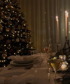 a table set for two with wine glasses and plates in front of a christmas tree