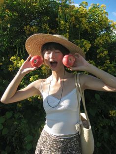 a woman wearing a straw hat holding two apples in front of her face with both hands