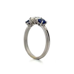 This exquisite three-stone engagement ring features a .93 carat antique cushion-cut diamond with VS2 clarity and J color. Flanked by two vibrant round blue sapphires, each weighing .46 carats, the sapphires add elegance to the overall design. The polished platinum band showcases intricate milgrain detailing. Decorative baskets cradle each gemstone, ensuring beauty from every angle. This ring blends refined craftsmanship with eye-catching details, making it a stunning choice. Luxury Three Stone Sapphire Ring, Three Stone Sapphire Round Ring Fine Jewelry, Timeless Round Three Stone Sapphire Ring, Luxury Three Stone Round Cut Sapphire Ring, Heirloom Three Stone Sapphire Ring, Heirloom Sapphire Three Stone Ring, Heirloom Sapphire Ring With Three Stones, Luxury Three-stone Sapphire Promise Ring, Luxury Three Stone Sapphire Promise Ring