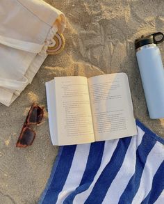 an open book and sunglasses are laying on the sand next to a water bottle, towel, and sunshade