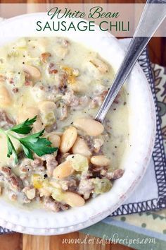 a white bowl filled with beans and meat soup on top of a wooden table next to a spoon