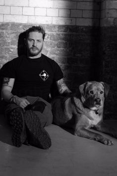 a man sitting next to a dog on top of a bed in front of a brick wall