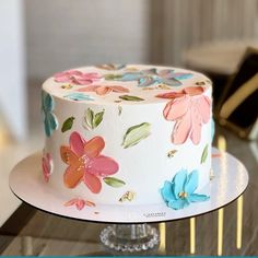 a white cake with colorful flowers on it sitting on top of a glass table next to a gold bar