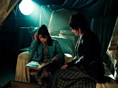 two women sitting on a bed in a room with blue drapes and curtains, one reading a book