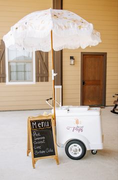 an ice cream cart with a white umbrella and menu board on the front door, in front of a house