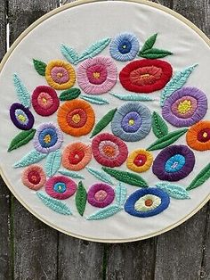 a white embroidery hoop with colorful flowers on it and leaves in the center, hanging on a wooden fence