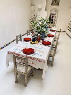 a long table with red plates and place settings on it in a white room filled with flowers