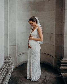 a pregnant woman in a white dress standing next to a wall and looking down at her belly