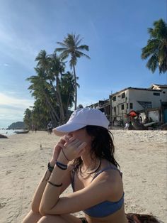 a woman sitting on top of a sandy beach next to palm trees and building in the background