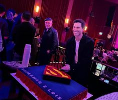 a man standing next to a giant superman cake in a room filled with other people