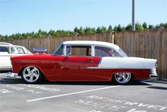 an old red and white car parked in a parking lot next to a wooden fence