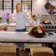 a woman standing in a kitchen next to a counter with fruit on it and two chairs