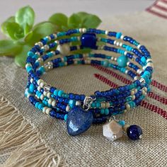 "Notice how the glass seed beads sparkle and shimmer in the video clip! And, I love these beautiful shades of blue all mixed with an accent of creamy beige in a type of bracelet that I wear quite often - memory wire!  Both the sparkle and matte finish of the seed beads add texture to this 5-wrap bracelet that is enhanced by the three focal beads - a tumbled Lapis nugget, a soft Riverstone round, and a lovely \"Goldline\" Turquoise (dyed Magnesite). The seed beads include a rich royal blue, soft matte turquoise, silver lined teal, and exquisite Duracoat matte galvanized silver.  The Swarovski crystal bi-cone beads include \"Golden Shadow\", \"Dark Sapphire\", and turquoise.  A few smaller Goldline Turquoise rounds are scattered throughout the bracelet as well.  The charms at one end of the Turquoise Bracelets With Heart Beads For Jewelry Making, Turquoise Bracelets With Heart And Round Beads, Turquoise Heart-shaped Beaded Bracelet, Adjustable Turquoise Beaded Bracelets With Heart Beads, Turquoise Beaded Bracelets With Heart Beads, Adjustable Blue Beaded Bracelets With Heart Beads, Turquoise Heart-shaped Beaded Bracelets, Blue Heart Beaded Bracelet With Round Beads, Blue Beaded Heart Bracelet