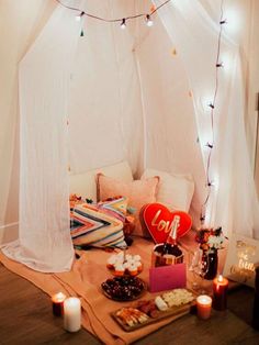 a bed covered in white curtains next to candles and other items on a table with a heart shaped pillow