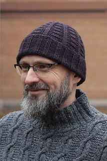 a bearded man wearing glasses and a knitted hat looks off to the side in front of a brick building