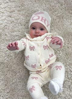 a baby is laying on the floor wearing a white hat and sweater with pink bows