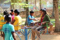 a group of people sitting around a blue table in the middle of a park with trees