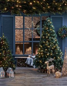 christmas trees and decorations in front of a window with lights on them, next to a fireplace