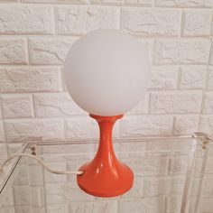 an orange table lamp with a white glass ball on it's base, against a brick wall