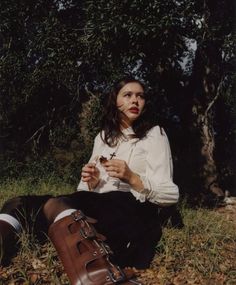 a woman sitting on the ground next to a tree with her hands in her pockets