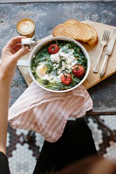 a bowl of food with eggs, tomatoes and bread on a wooden cutting board next to a cup of coffee