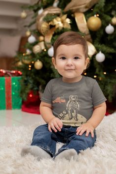 a baby sitting in front of a christmas tree wearing a t - shirt with a cactus on it