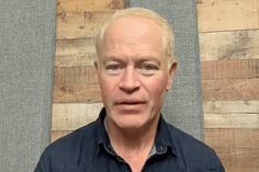 a man in a blue shirt standing next to a wooden wall and looking at the camera