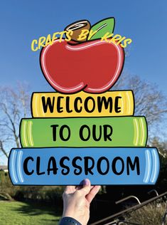 a hand holding up a sign that says welcome to our classroom on top of books