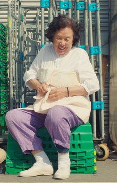 a woman sitting on top of a green crate next to stacks of plastic crates and holding a cup