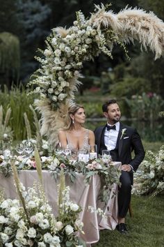 a man and woman sitting at a table in front of an arch decorated with flowers