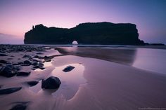 the beach is covered in rocks and water at dusk, with an arch shaped rock sticking out of the sand