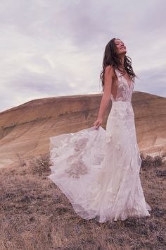 a woman in a white dress standing on top of a dry grass covered field next to a hill