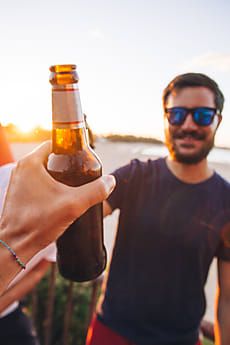 a man holding up a beer bottle in front of another man by the ocean at sunset