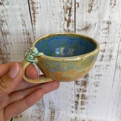 a hand holding a blue and gold cup with a frog on the handle, against a white wooden background