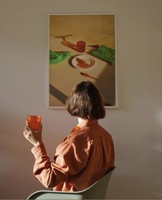 a woman sitting in a chair holding a cup and looking at a painting on the wall