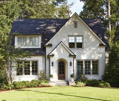 a white house with black shingles and trees in the front yard, surrounded by lush green grass