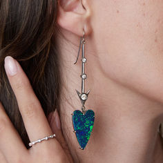 A close-up image showcasing an elegant opal earring by Sylva & Cie. The design features a vibrant heart-shaped opal in shades of blue and green, set in a sleek silver frame. Above the opal, a delicate vertical bar is adorned with sparkling diamonds, adding a touch of sophistication. The earring is styled on a model, highlighting its bold and luxurious aesthetic. Perfect for making a statement, this piece reflects Sylva & Cie’s signature craftsmanship. Opal Earring, European Cut Diamonds, Opal Earrings, Heart With Arrow, Opal Jewelry, Jewelry Lover, Diamond Heart, Statement Earrings, Heart Shapes