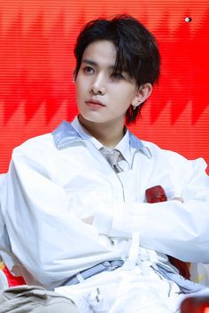 a young man sitting in front of a red wall wearing a white shirt and tie