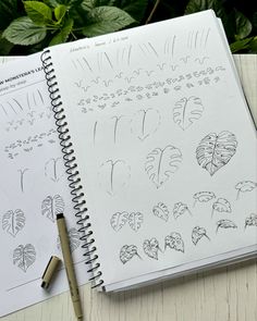 an open notebook with drawings on it next to a pen and some plants in the background